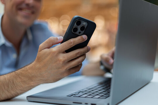 smiling man using a smartphone while working on his laptop. Close-up of hands holding the phone at a white desk with blurred warm bokeh background. Perfect for freelance and modern technology.