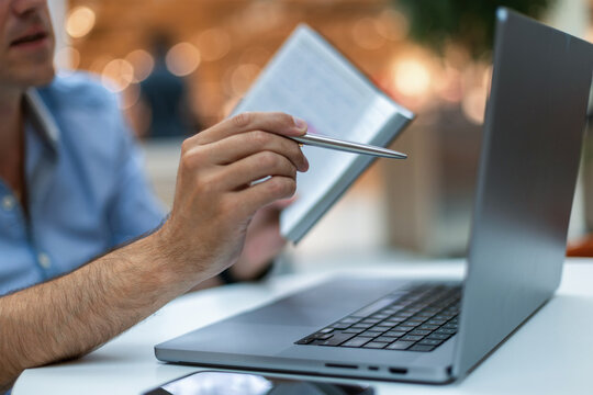 Close-up of a man's hands as he uses a pen to point at a laptop screen while holding a notebook. Focus on data analysis, remote work, or business strategy planning.