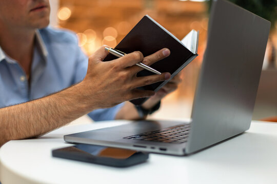 Close-up of a man's hands holding an open notebook and a pen while working on a laptop computer. Focus on schedule planning, remote business, and education with warm bokeh background lights.