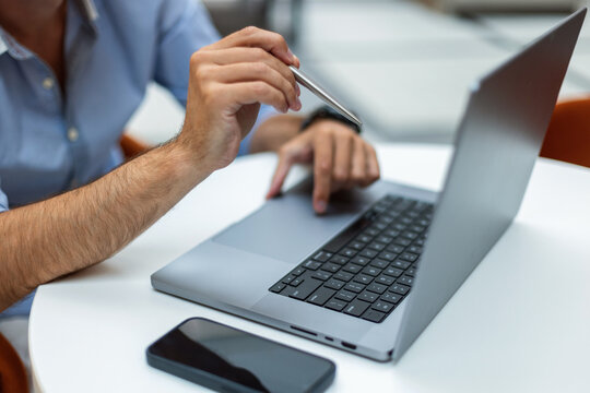 Close-up of a businessman's hands pointing at a laptop screen with a silver pen while working at a white desk. Smartphone in the foreground. Perfect for technology and remote work concepts.
