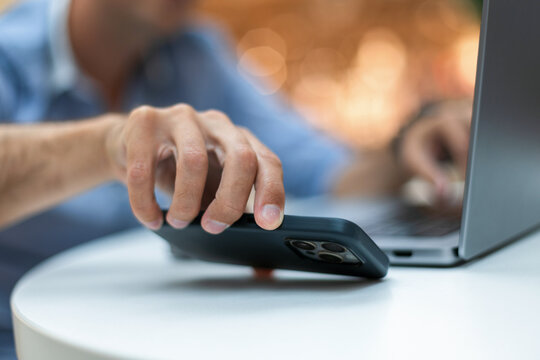 Close-up of a man's hand reaching for a smartphone on a desk while working on a laptop computer. Blurred warm bokeh background. Perfect for technology, multitasking, and remote work concepts.