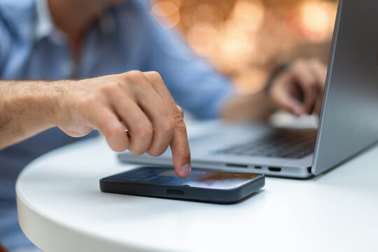 Focused shot of male hands multitasking. Right index finger touches a smartphone screen while left hand works on a laptop. businessman taps on a notification on his smartphone