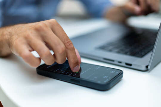 Close-up of a man's hand using a smartphone with an application on screen, while working on a laptop at a white desk. Perfect for technology, finance, mobile banking, and remote work concepts.