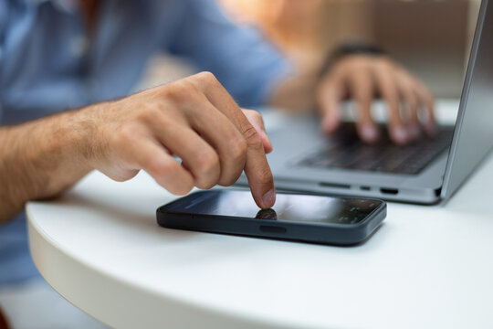Close-up of a man's hand touching a smartphone screen while typing on a laptop computer. Blurred background, white desk. Great for technology, multitasking, and modern remote business concepts.