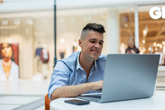 A smiling young man working on a laptop at a table in a modern public space or mall. A smartphone lies on the desk. Ideal for freelance, remote business, and digital nomad lifestyle concepts.
