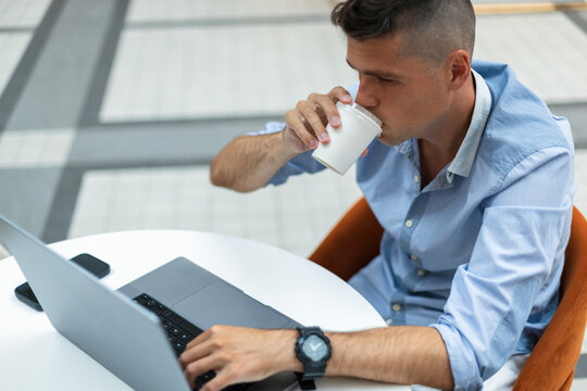 young businessman drinking coffee from a paper cup while working on a laptop at a white table. Ideal for freelance, remote work, and digital nomad lifestyle concepts.