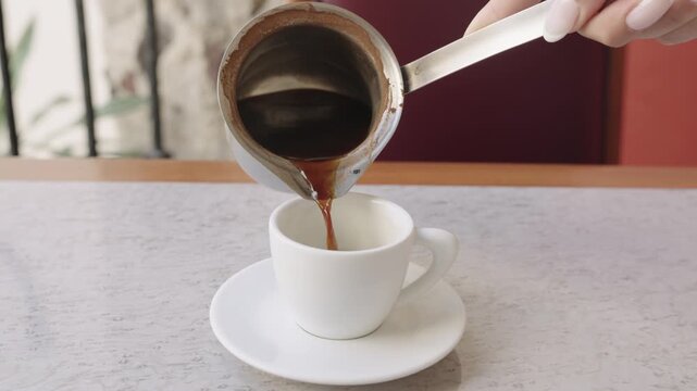 Woman's hand pouring hot black coffee from a cezve into a white ceramic cup on a cafe table