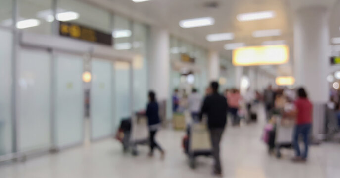 A blurry image of a busy airport terminal with travelers and luggage carts. The scene captures people moving through the space amid various airport signage.