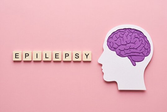 A conceptual representation of epilepsy featuring a paper-cut human head silhouette with a brain and the word EPILEPSY spelled out in Scrabble-style tiles on a soft pink background.
