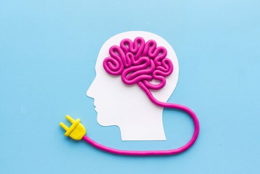 A conceptual representation of a human head silhouette with a pink clay brain plugged into a yellow electrical outlet, symbolizing creative energy and artificial intelligence.