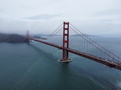 Aerial view of the iconic Golden Gate Bridge spanning the bay with traffic crossing under a cloudy sky and fog rolling over the coastal hills in San Francisco, California, United States.