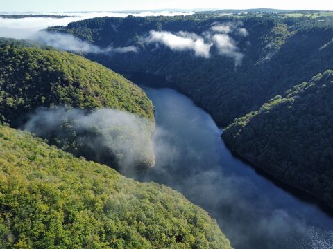 Aerial view of the Dordogne River winding through lush green forested hills with low-hanging morning mist in Argentat-sur-Dordogne, Nouvelle-Aquitaine, France.