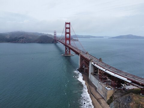 Aerial view of the Golden Gate Bridge spanning the bay with traffic on the deck, rocky coastline, and overcast sky in San Francisco, California, United States.