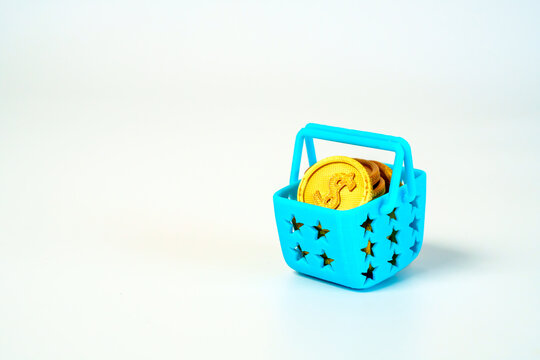 Small blue plastic shopping basket filled with golden dollar coins on a white background. Representing retail, money, savings, wealth, and budget. Minimalist still life for financial concepts.