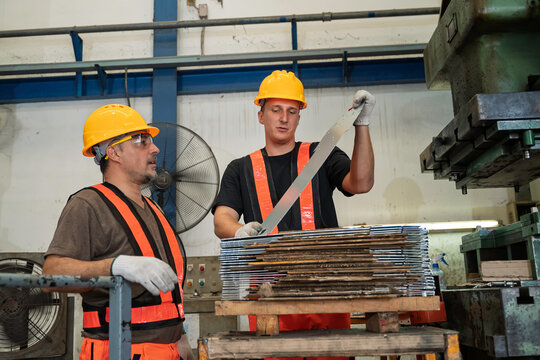 Two professional male industrial factory workers wearing yellow safety helmets carefully inspect a steel sheet metal part near a heavy automated stamping press machine during production.