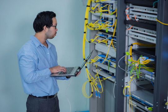 Focused Asian male IT network administrator uses a laptop to monitor server performance inside a bright data center managing complex corporate cybersecurity operations and infrastructure.