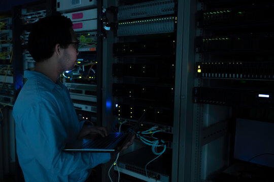 Focused Asian male IT network administrator uses a laptop to monitor server performance inside a dark data center room managing complex corporate cybersecurity operations and infrastructure.