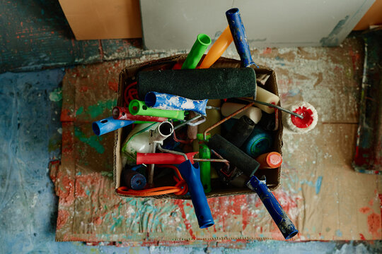 Top view shot of paint rollers and handles filling cardboard box on splattered studio floor, suggesting contemporary artist workflow. Useful for creative business, renovation, art process marketing