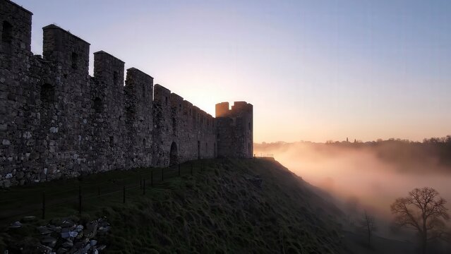 Ancient stone castle wall with towers illuminated by sunrise, surrounded by misty landscape and grassy hill, showcasing historical architecture in serene setting