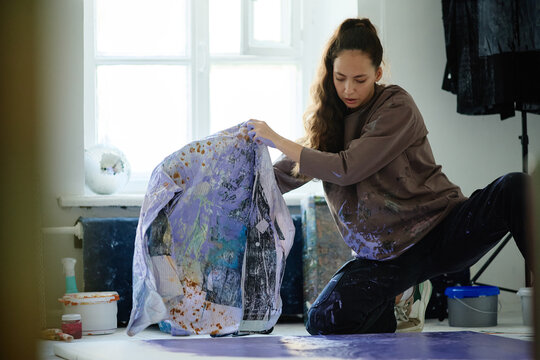 Young adult Caucasian woman creating contemporary artwork in studio, handling paint covered fabric and canvas. Ideal for creative industry marketing, artist branding, workshop promotion