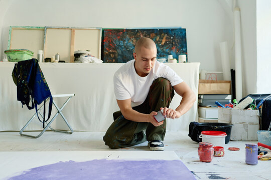 Young adult Caucasian man creating contemporary artwork in studio, opening paint jar while preparing canvas. Ideal for creative business, artist branding, art marketing