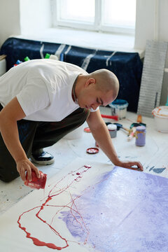 Vertical shot of young adult Caucasian man creating abstract painting on canvas floor, pouring paint in art studio. Supporting contemporary art marketing, creative business branding