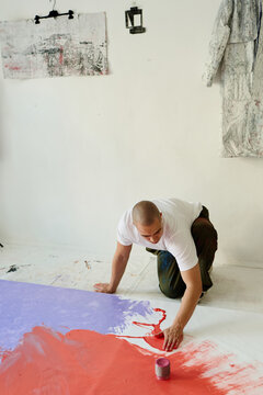 Vertical shot of young adult Caucasian man creating abstract artwork on floor canvas, spreading paint by hand in studio. Ideal for contemporary art, creative process, artist branding