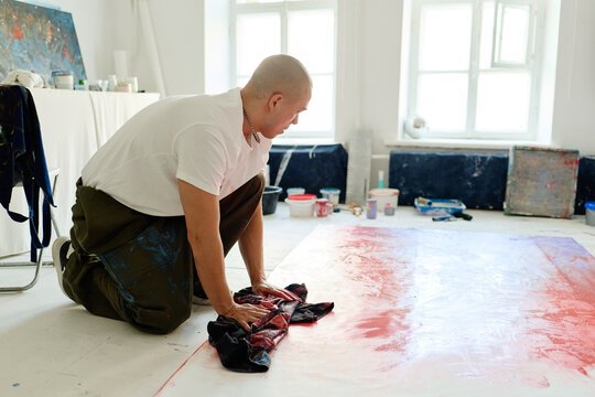 Young adult Caucasian man kneeling and wiping large canvas on studio floor, creating contemporary artwork and creative process concept. For editorial, culture, artist branding, gallery promotion