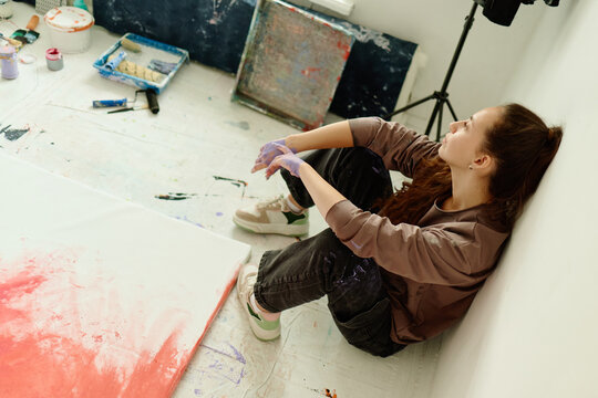 Young adult Caucasian woman resting beside canvas in art studio, reflecting after painting session and developing creative process. Ideal for contemporary artist branding and art education