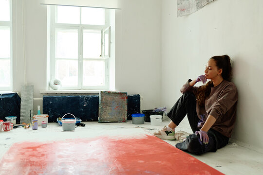 Young adult Caucasian woman artist resting beside large canvas in studio, contemplating creative process and artistic work. Useful for contemporary art, creative burnout, inspiration marketing