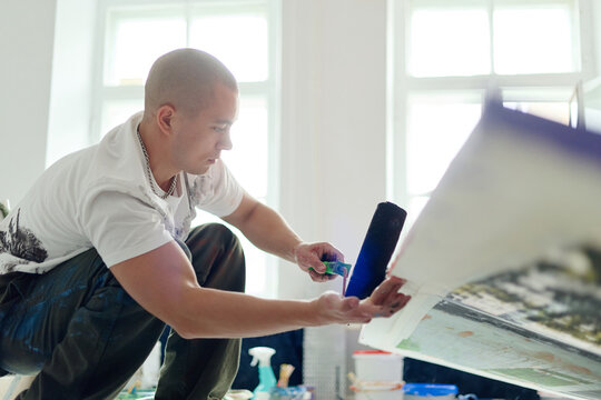 Young adult Caucasian male artist applying ink with roller on large print in studio, creating contemporary artwork. Useful for creative business, art production, design marketing