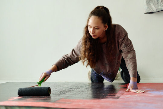 Young adult Caucasian woman painting canvas on floor with roller in artist studio, creating contemporary artwork. Ideal for creative process, contemporary art, maker branding campaigns