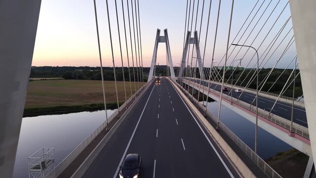 Aerial view of cars traveling on highway bridge at dusk. Continuous flow of vehicles over scenic landscape with fields and water visible in background. Autos driving at speed road. Cars rides at route