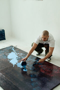 Vertical shot of young adult man pouring paint onto large canvas and spreading pigment on studio floor, creating contemporary artwork. Ideal for creative industry branding and artist promotion