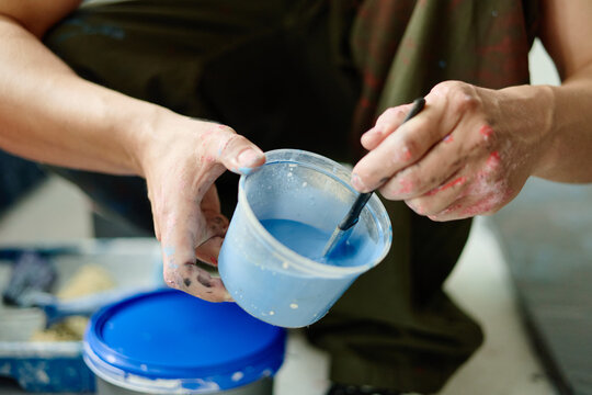 Close up of contemporary artist hands mixing blue paint in plastic cup, preparing materials for artwork production. Useful for creative process, studio practice, art workshop marketing