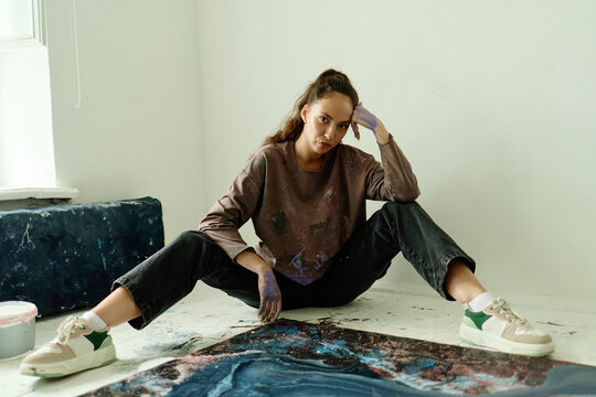 Portrait of young adult Caucasian woman artist sitting on studio floor beside abstract canvas, pausing after painting. Ideal for creative profession, art process, maker branding
