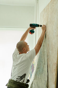 Vertical shot of young adult Caucasian man mounting large artwork on wall, using power drill in studio. Suitable for contemporary art, creative workspace, installation service marketing
