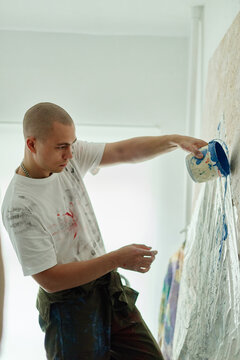 Vertical shot of young adult Caucasian man pouring blue paint onto large canvas, creating contemporary artwork in studio. Ideal for creative industry marketing, artist branding