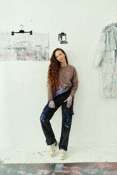 Vertical portrait of young adult Caucasian woman standing in art studio, showing paint stained hands and clothing, representing contemporary artist branding. For creative business promotion