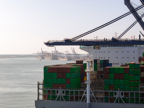 Aerial view of a large container ship loaded with colorful shipping containers docked at a busy industrial port with cranes in the background United Kingdom.