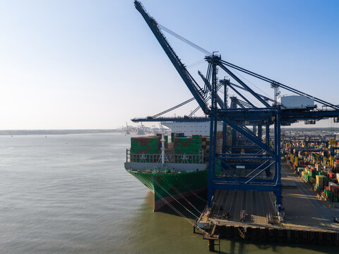 Aerial view of a large container ship docked at a port with industrial cranes and stacked shipping containers in the United Kingdom.