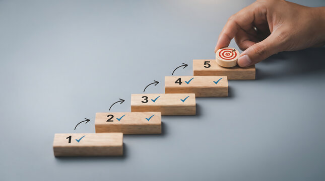 Hand placing wooden block with target icon on step 5, stairs concept with check marks on blocks 1 to 4, achieving goals and planning business strategy.