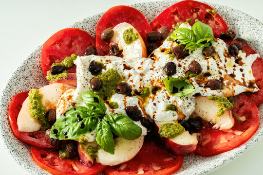 Close up of Italian style salad with tomatoes, peaches, olives, burrata and pesto on white background
