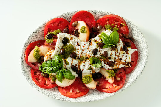 Italian style salad with tomatoes, peaches, olives, burrata and pesto on white background