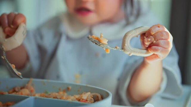 Close up hands of little Asian toddler holding spoon and fork eating food at home high chair, messy cute kid self feeding for baby led weaning BLW, early childhood development, nutrition and parenting