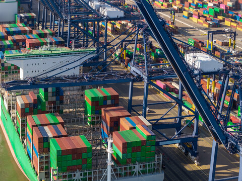 Aerial view of a large container ship being loaded with colorful shipping containers by massive gantry cranes at a busy industrial port in the United Kingdom.