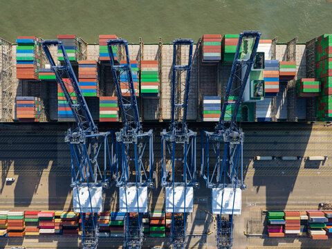 Aerial view of a large container ship being loaded with colorful shipping containers by massive blue gantry cranes at a busy port in the United Kingdom.