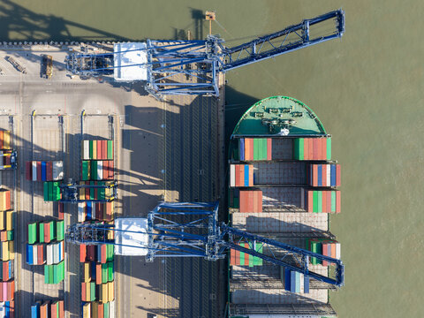 Aerial view of a large container ship being loaded with colorful shipping containers by industrial cranes at a busy port terminal in the United Kingdom.