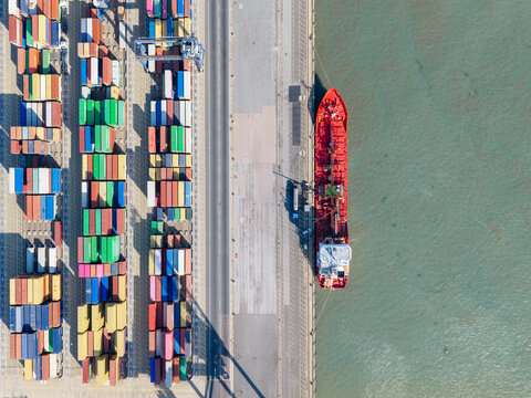 Aerial view of a red cargo ship docked at a busy container terminal with rows of colorful shipping containers stacked on the wharf in the United Kingdom.