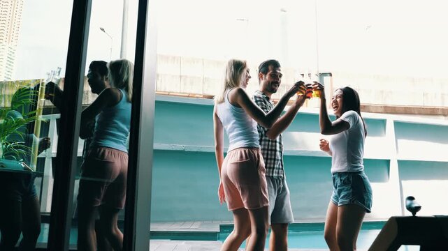 Group of multiethnic friends dancing, drinking beverages, toasting glass bottles laughing together by swimming pool with backlit sunlight. Happy men and women enjoying pool party, summer vacation 